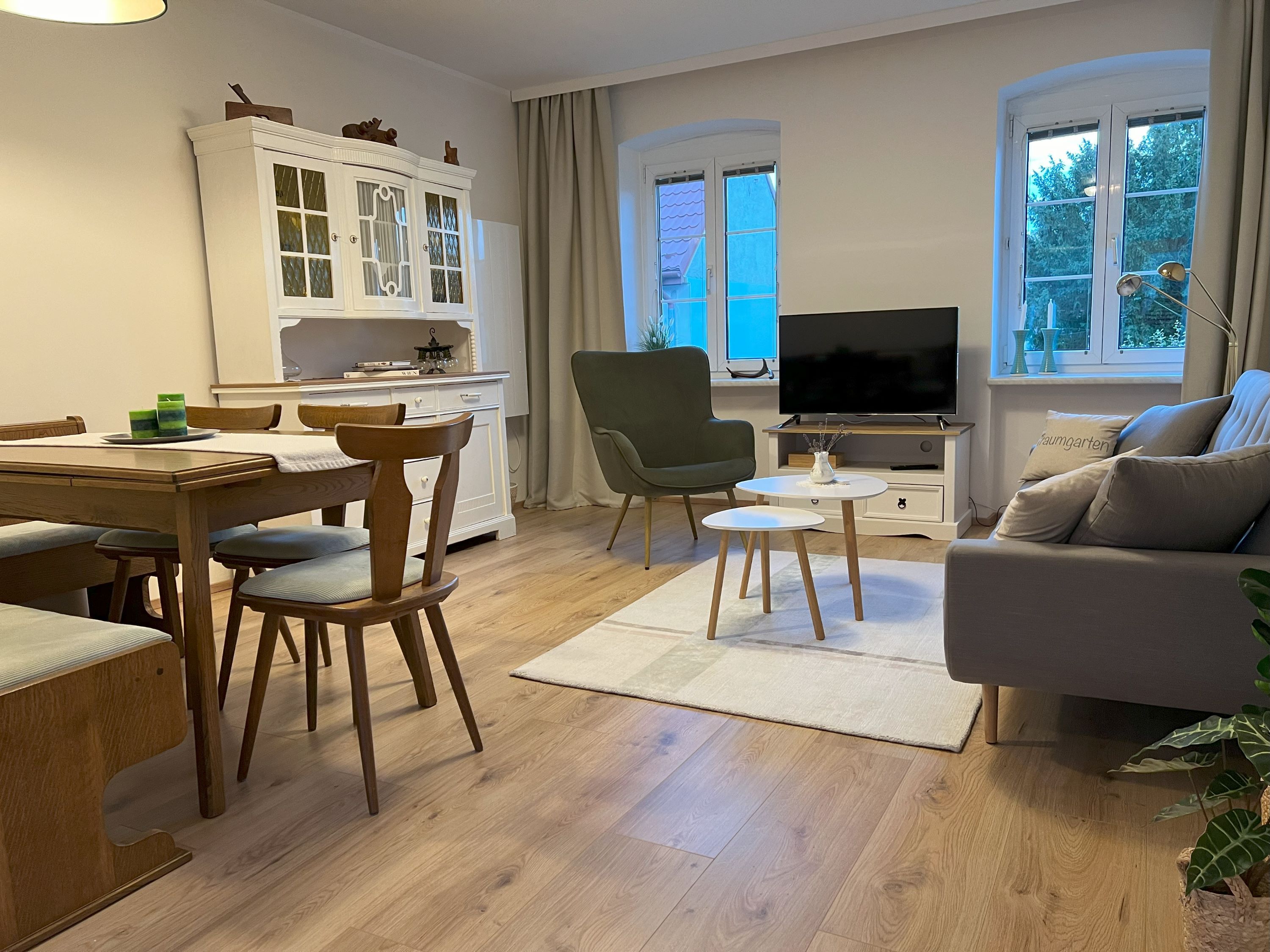 Gemütliches Wohnzimmer mit Holzmöbeln, Sofa, Sessel, Esstisch und Fernseher. Helle Wände und Holzboden.