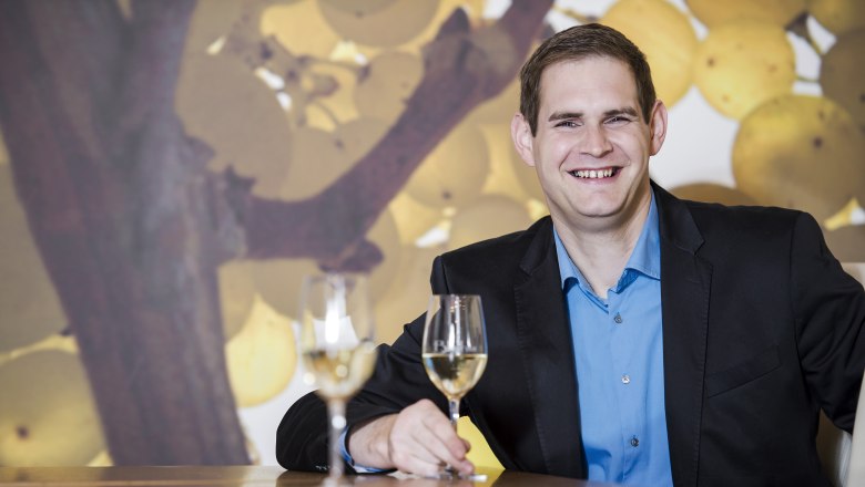 Ein Mann in einem Anzug sitzt an einem Tisch mit zwei Weingläsern vor einem Hintergrund mit Weintrauben.