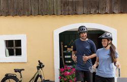 Zwei Radfahrer genießen eine entspannte Pause vor einem charmanten, gelb gestrichenen Haus, umgeben von blühenden Pflanzen. Die frische Luft und die malerische Landschaft des Weinviertels laden dazu ein, die Umgebung zu erkunden und die Schönheit der Natur zu erleben.