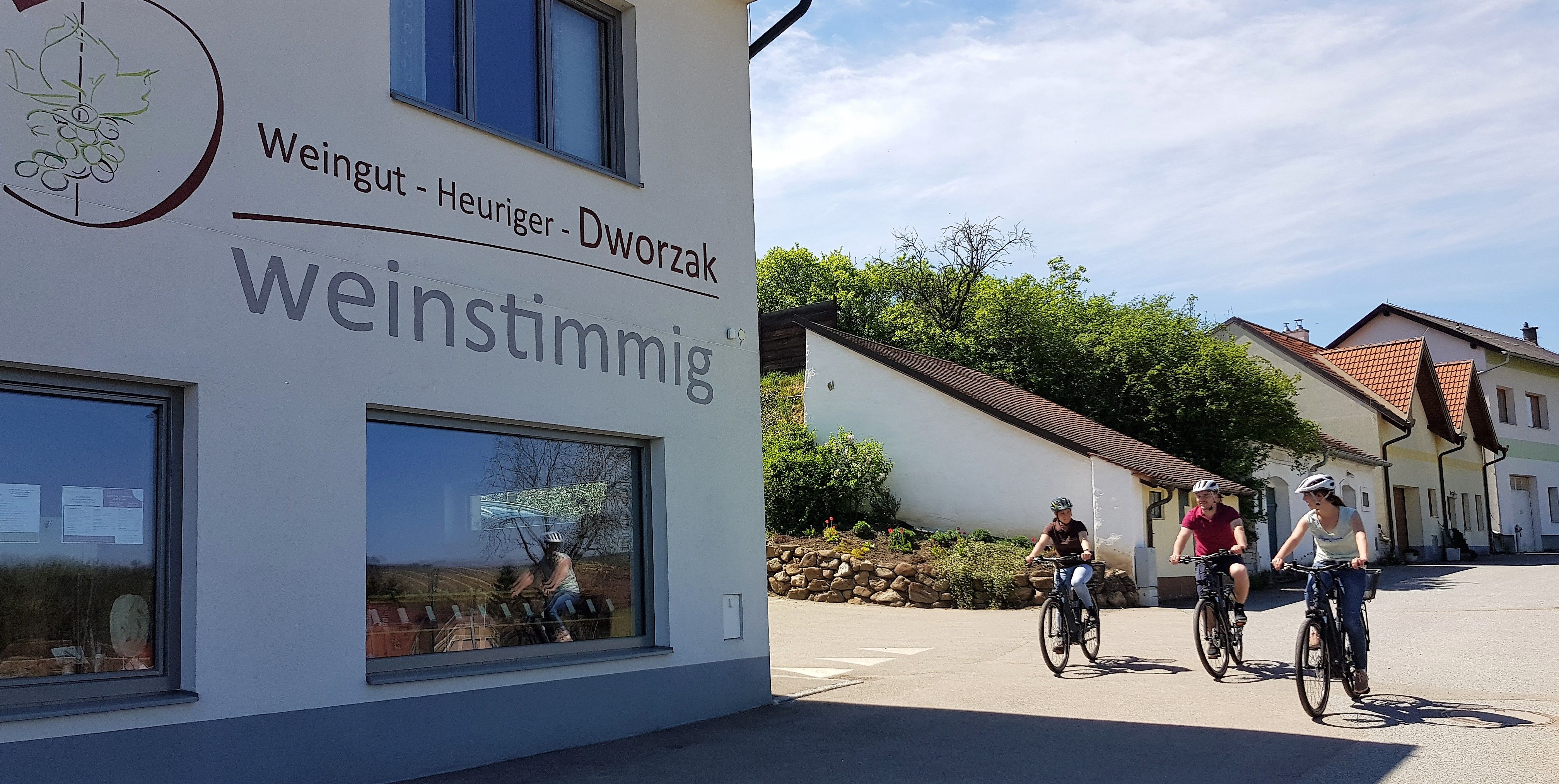 Drei Personen fahren mit Fahrrädern an einem Gebäude mit der Aufschrift 'Weingut - Heuriger - Dworzak weinstimmig' vorbei.