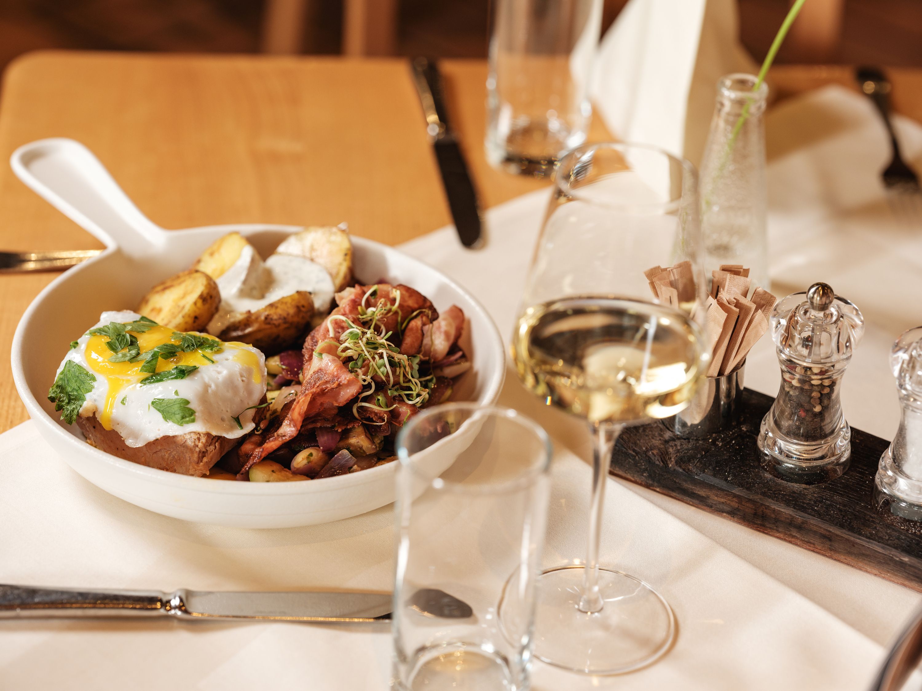 Ein Teller mit Fleisch, Spiegelei, Kartoffeln und Gemüse, serviert mit einem Glas Weißwein auf einem gedeckten Tisch.