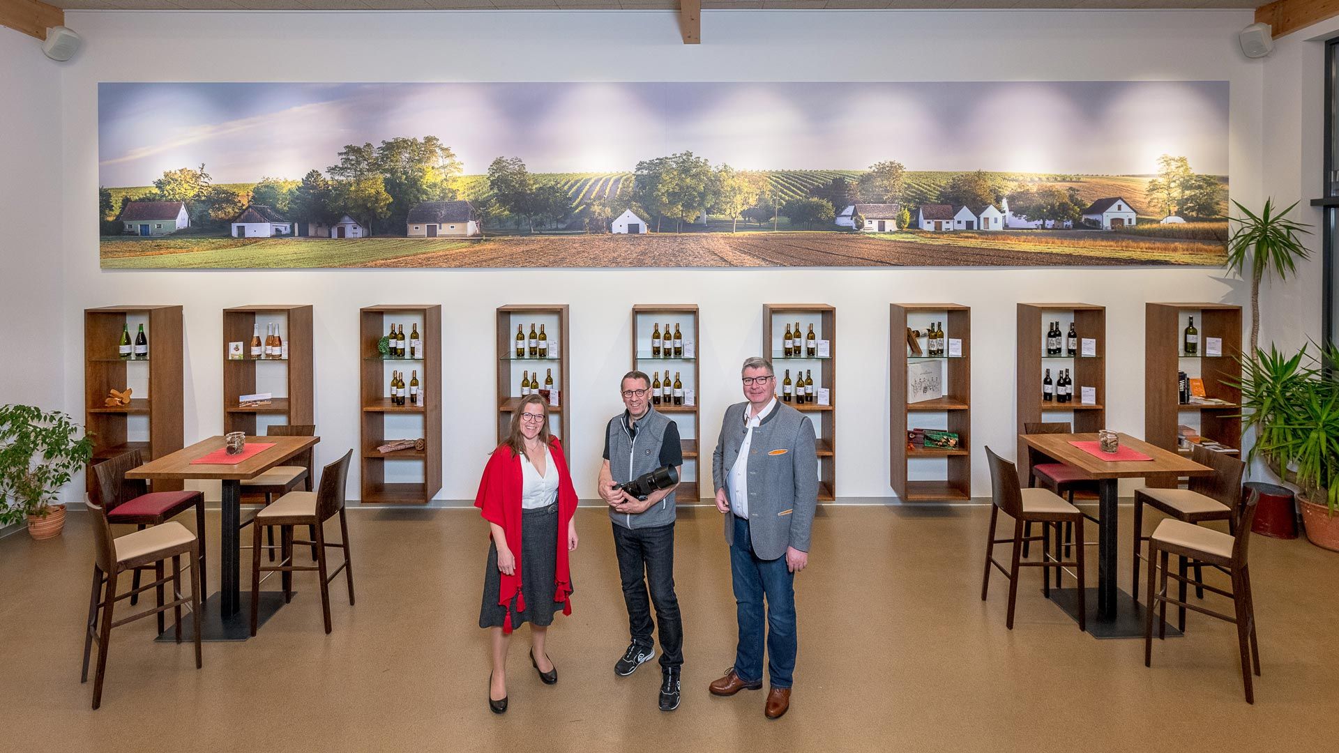 Drei Personen stehen in einem Weingut vor Regalen mit Weinflaschen und einem großen Wandbild von Weinbergen.