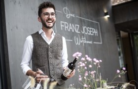Mann mit Brille und Weste hält Weinflasche vor Restaurant 'Franz Joseph'.