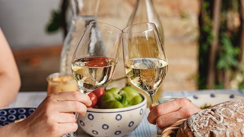 Two people toast with glasses of white wine, fruit and bread in the background.