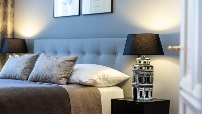 Elegantes Doppelzimmer mit grauem Kopfteil, zwei Kissen, Nachttischlampe und Wandbildern.