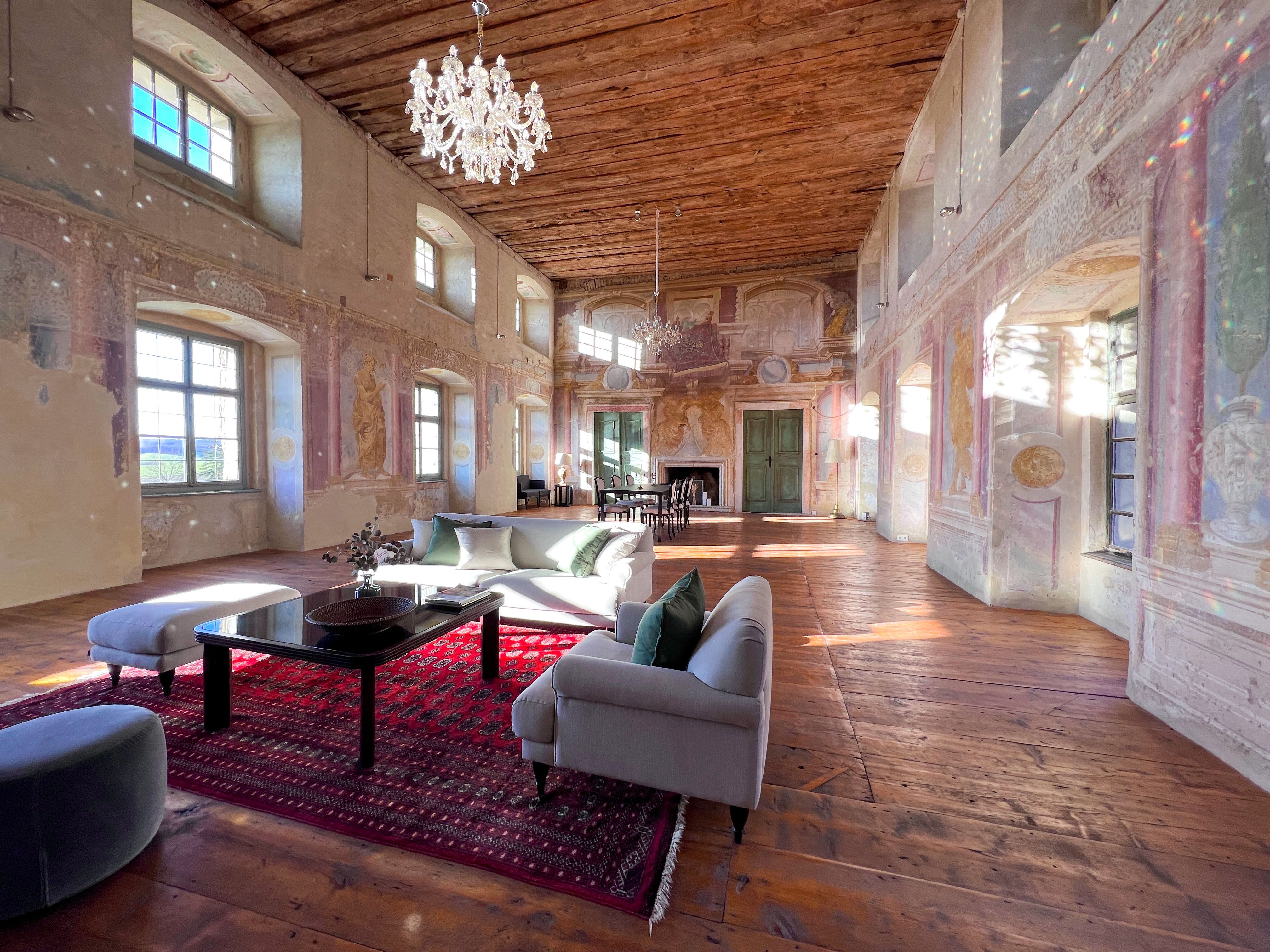 Ein historischer Festsaal mit Holzboden, bemalten Wänden und einem Kronleuchter. Moderne Möbel und ein roter Teppich sind im Raum platziert.