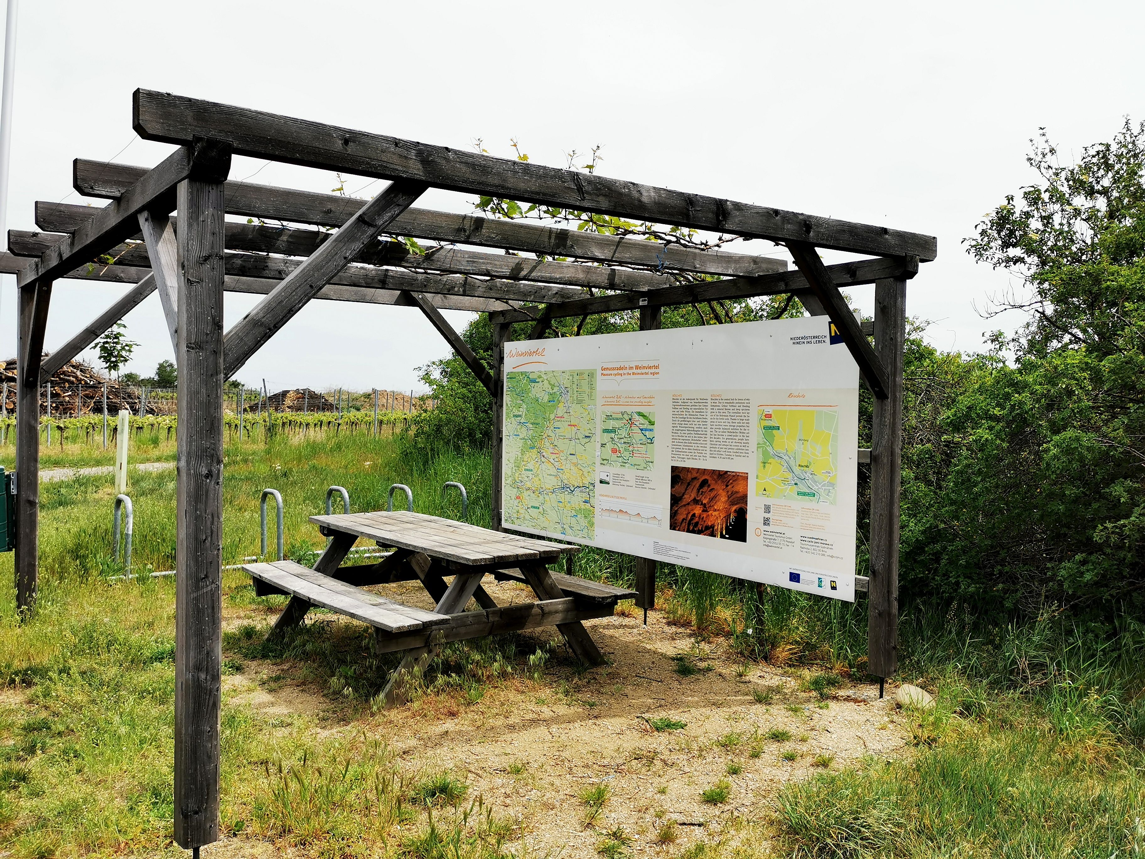 Holzpergola mit Infotafel und Picknicktisch in einer ländlichen Umgebung.
