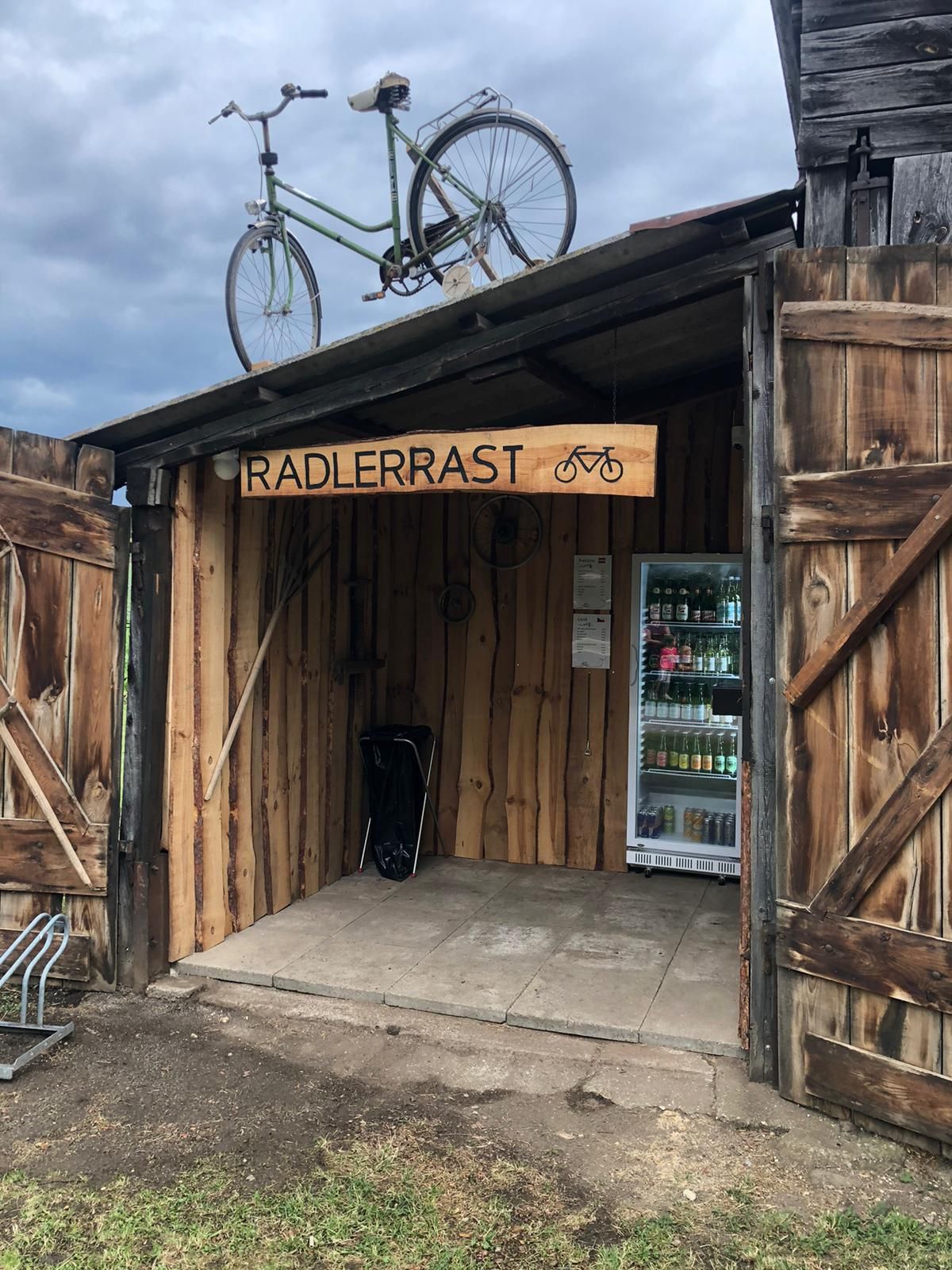 Ein hölzerner Unterstand mit einem Fahrrad auf dem Dach und einem Kühlschrank mit Getränken. Ein Schild mit der Aufschrift 'Radlerrast'.