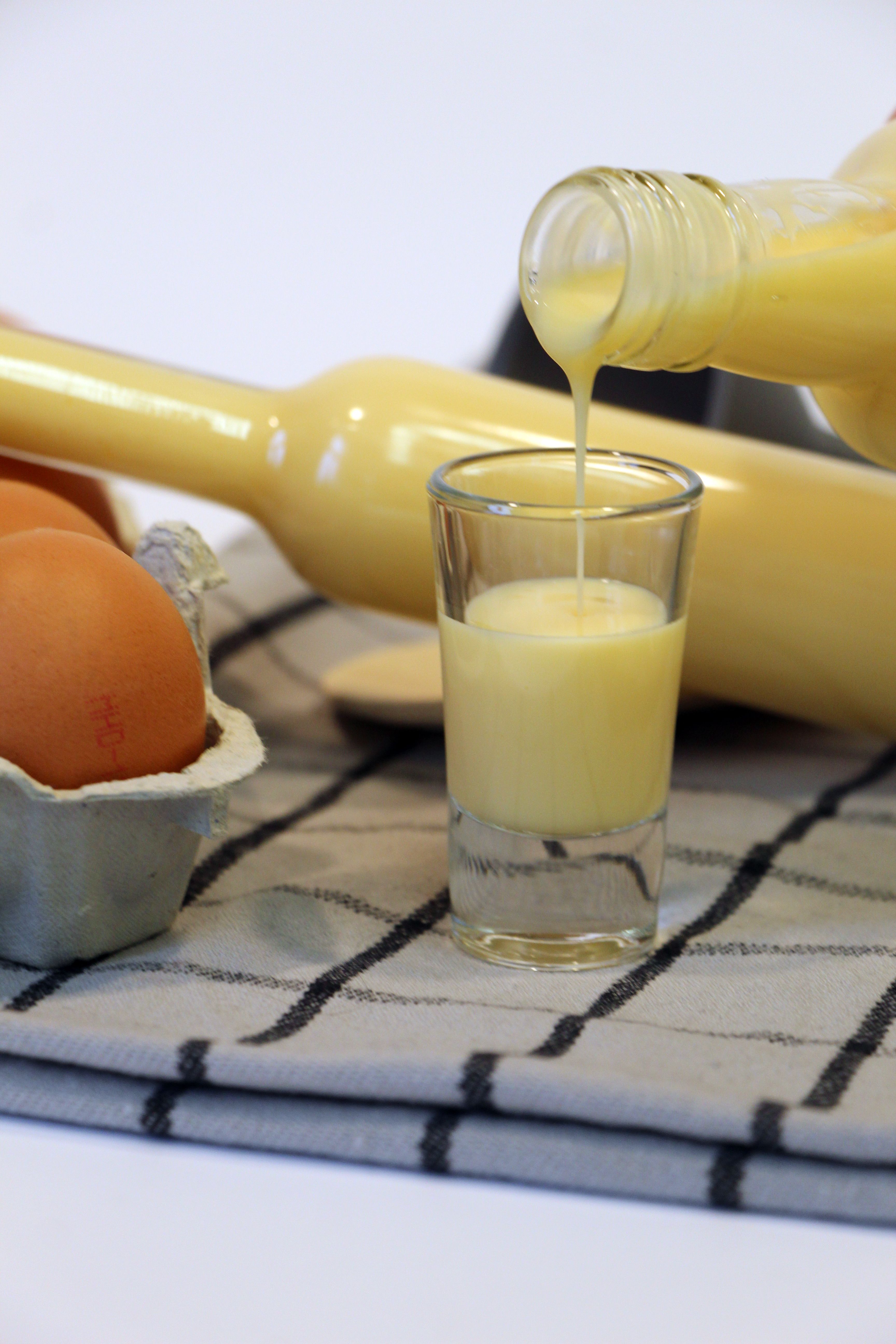 In ein Stamperl wird aus einer Glasflasche Eierlikör eingegossen, im Hintergrund sind Eier und eine weitere Flasche Eierlikör zu sehen.