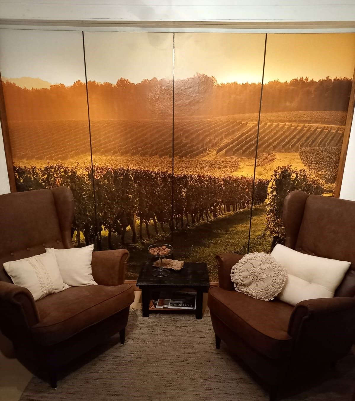 Zwei braune Sessel mit Kissen vor einer Wand mit Landschaftsfototapete eines Weinbergs.
