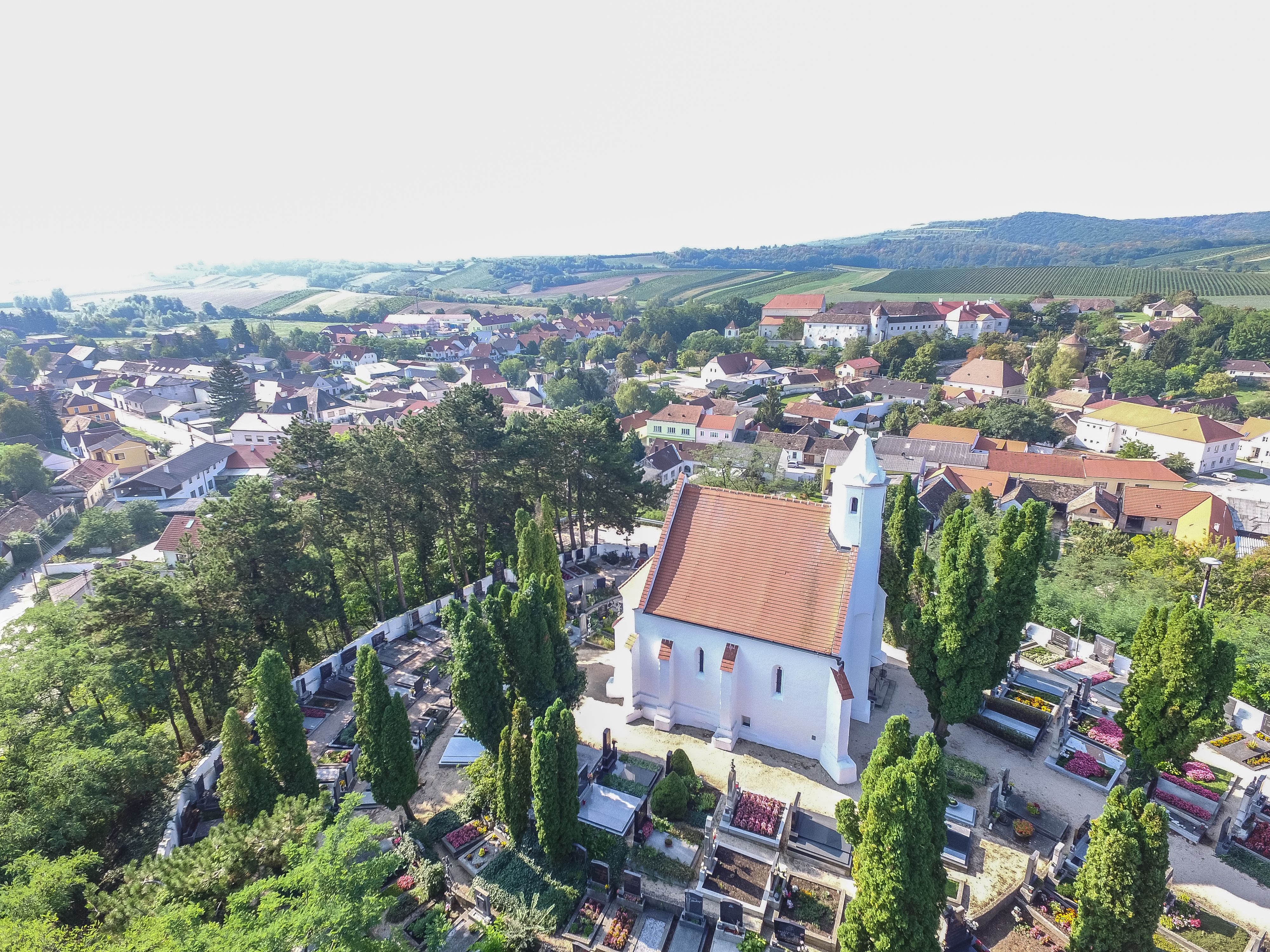 Luftaufnahme einer Kirche mit rotem Dach, umgeben von einem Friedhof und Bäumen, in einer ländlichen Umgebung mit Häusern und Hügeln im Hintergrund.
