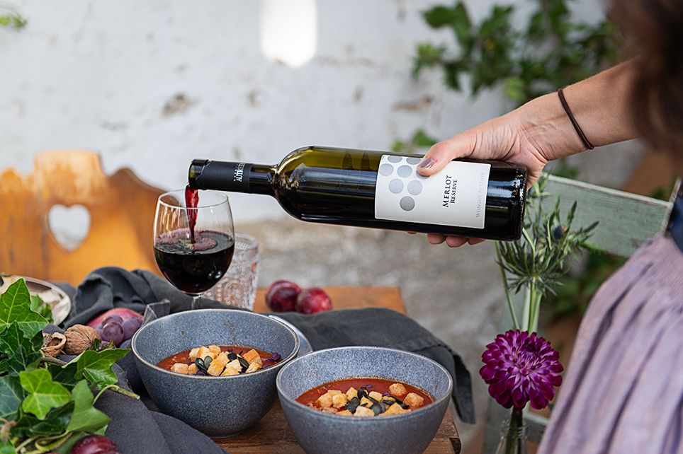 A hand pours red wine into a glass next to two bowls of soup on a table.