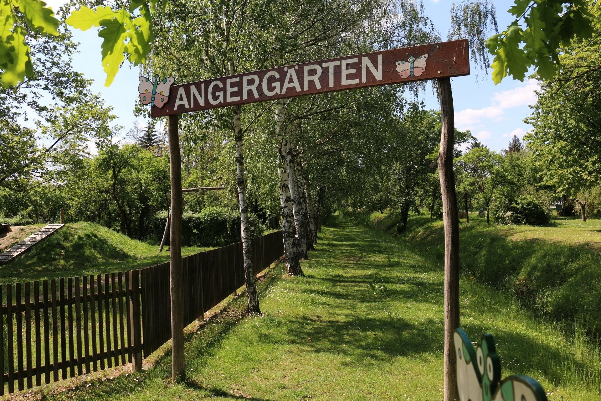 Eingang zu den Angergärten mit Holzschild und Bäumen im Hintergrund.