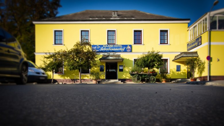 Gelbes Gebäude mit der Aufschrift 'Auland-Hotel Siebenbrunnerhof', umgeben von Bäumen und einem alten Wagen.