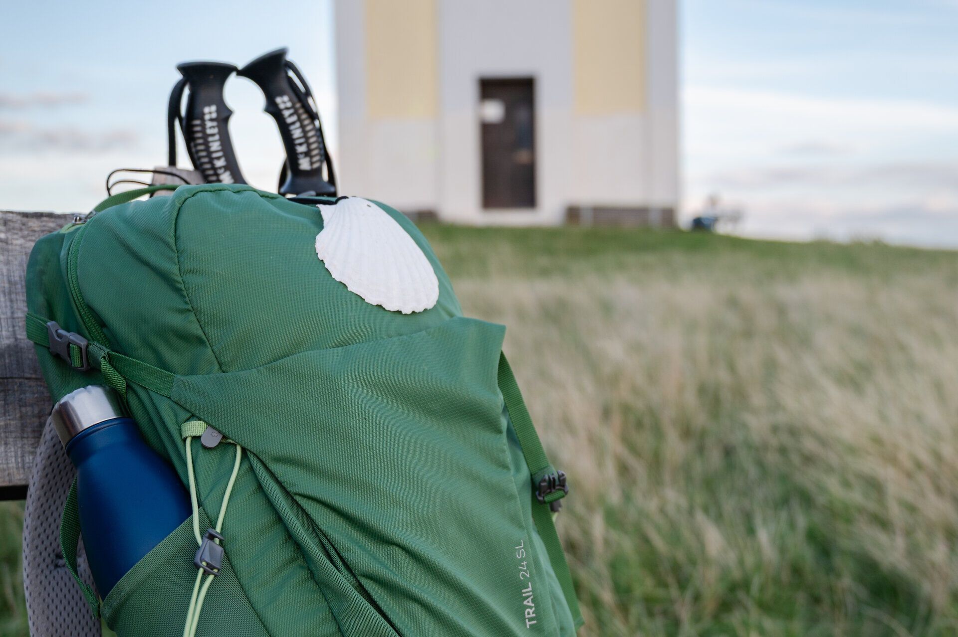 Ein Rucksack mit Jakobswegmuschel und Trinkflasche lehnt an einer Bank, dahinter ragen Wanderstöcke hervor.