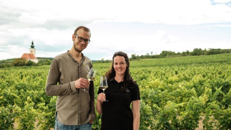 Two people stand with wine glasses in a vineyard, with a church in the background.