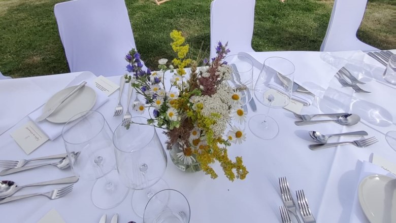 Gedeckter Tisch mit Blumenstrauß vor Weinberg und Liegestühlen.
