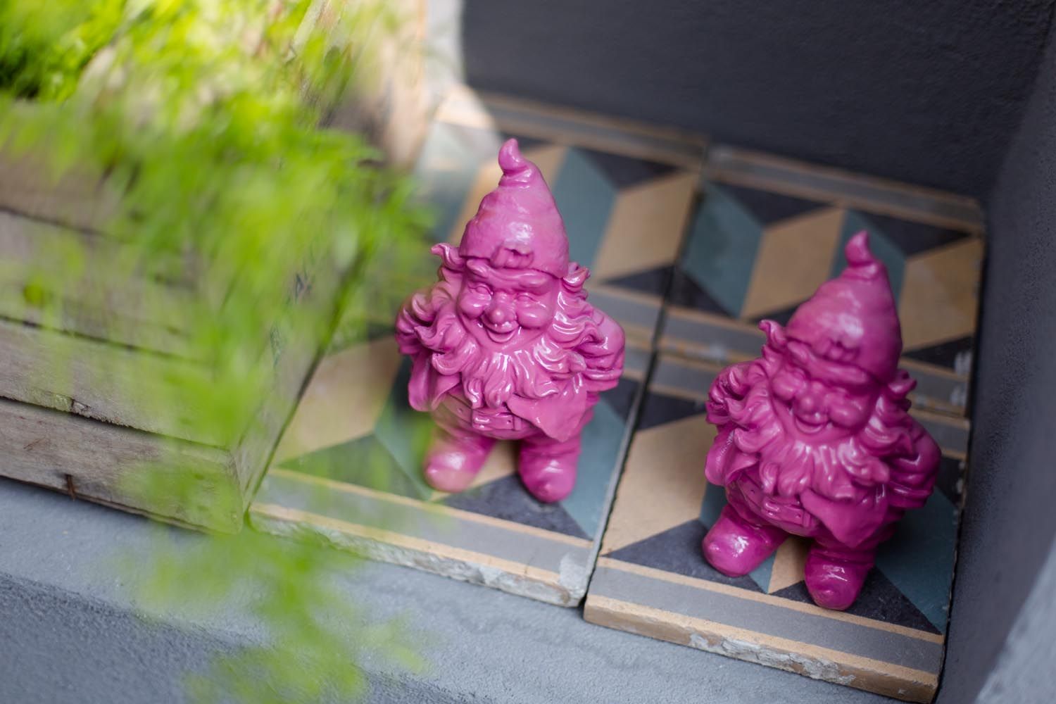 Zwei pinke Gartenzwerge auf gemusterten Fliesen neben einer Holzkiste mit Pflanzen.