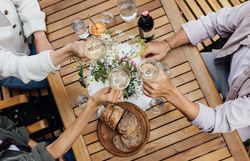 Inmitten einer idyllischen Atmosphäre stoßen Freunde mit Gläsern auf die schönen Momente des Lebens an. Der Tisch ist liebevoll gedeckt mit frischem Brot und einer bunten Auswahl an Köstlichkeiten, während die Blumen in der Mitte des Tisches die Freude des Augenblicks unterstreichen.