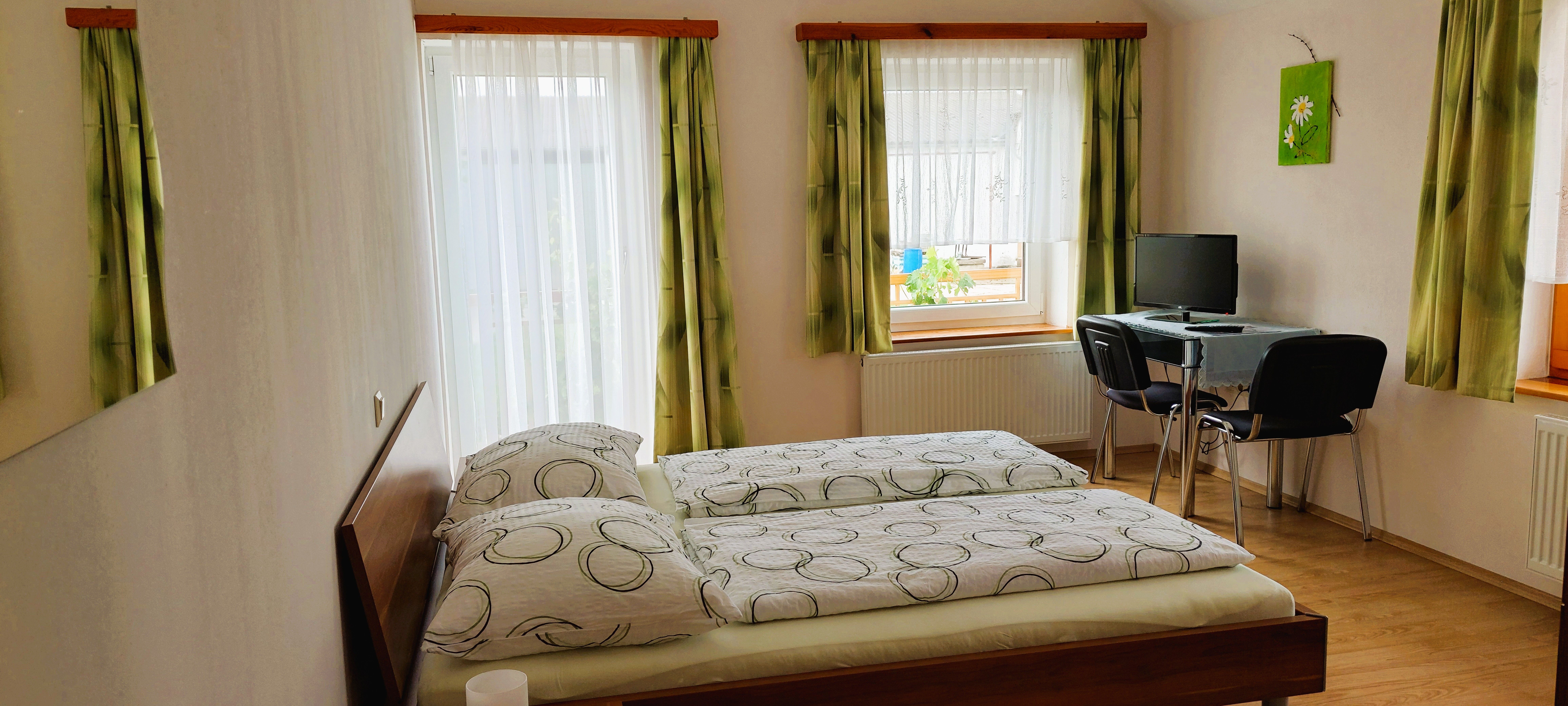 Ein helles Schlafzimmer mit zwei Einzelbetten, grün gemusterten Vorhängen und einem kleinen Schreibtisch mit Stuhl und Computer.