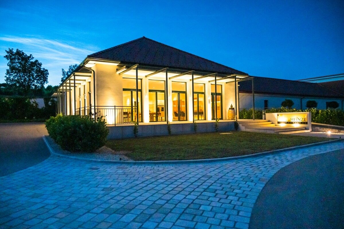 Ein modernes Gebäude mit Glasfronten, beleuchtet in der Dämmerung, umgeben von einem gepflasterten Weg und Grünflächen.