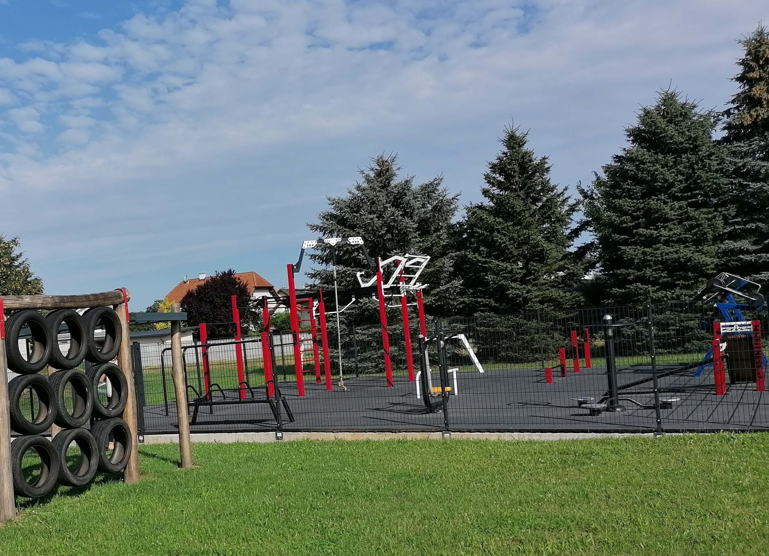 An outdoor fitness park with red and black equipment, surrounded by trees and a fence. In the foreground, tires are attached to wooden posts.