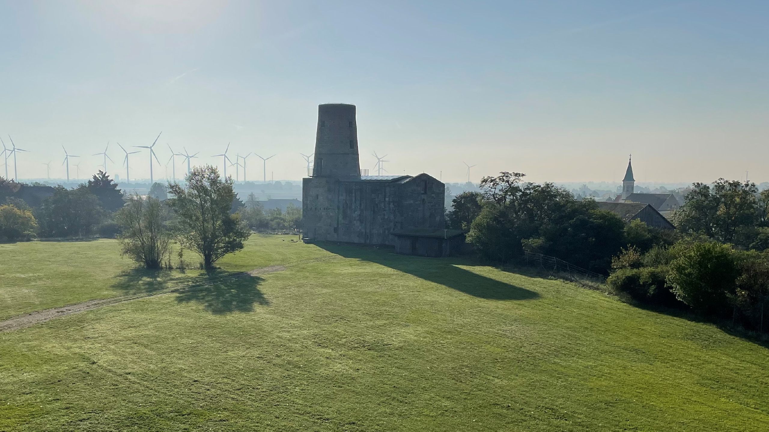 Aussicht Ruine: Weite grüne Wiese mit einer alten Ruine im Zentrum, im Hintergrund Windräder und rechts eine Kirchturmspitze.