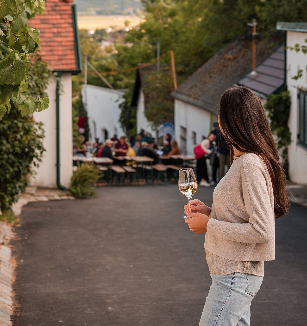 Eine Frau steht mit einem Glas Weißwein in der Hand in einer Kellergasse und betrachtet das gesellige Treiben, das im Rahmen des Kellergassenfestes herrscht.