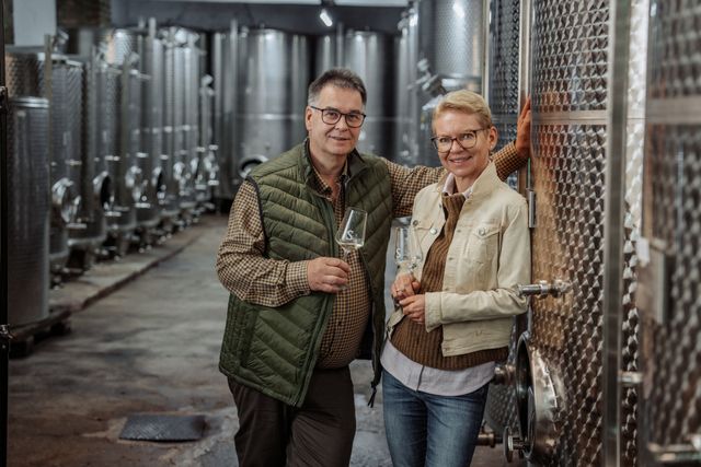 Ein Mann und eine Frau stehen in einer Weinkellerei vor großen Edelstahltanks, beide halten ein Weinglas.