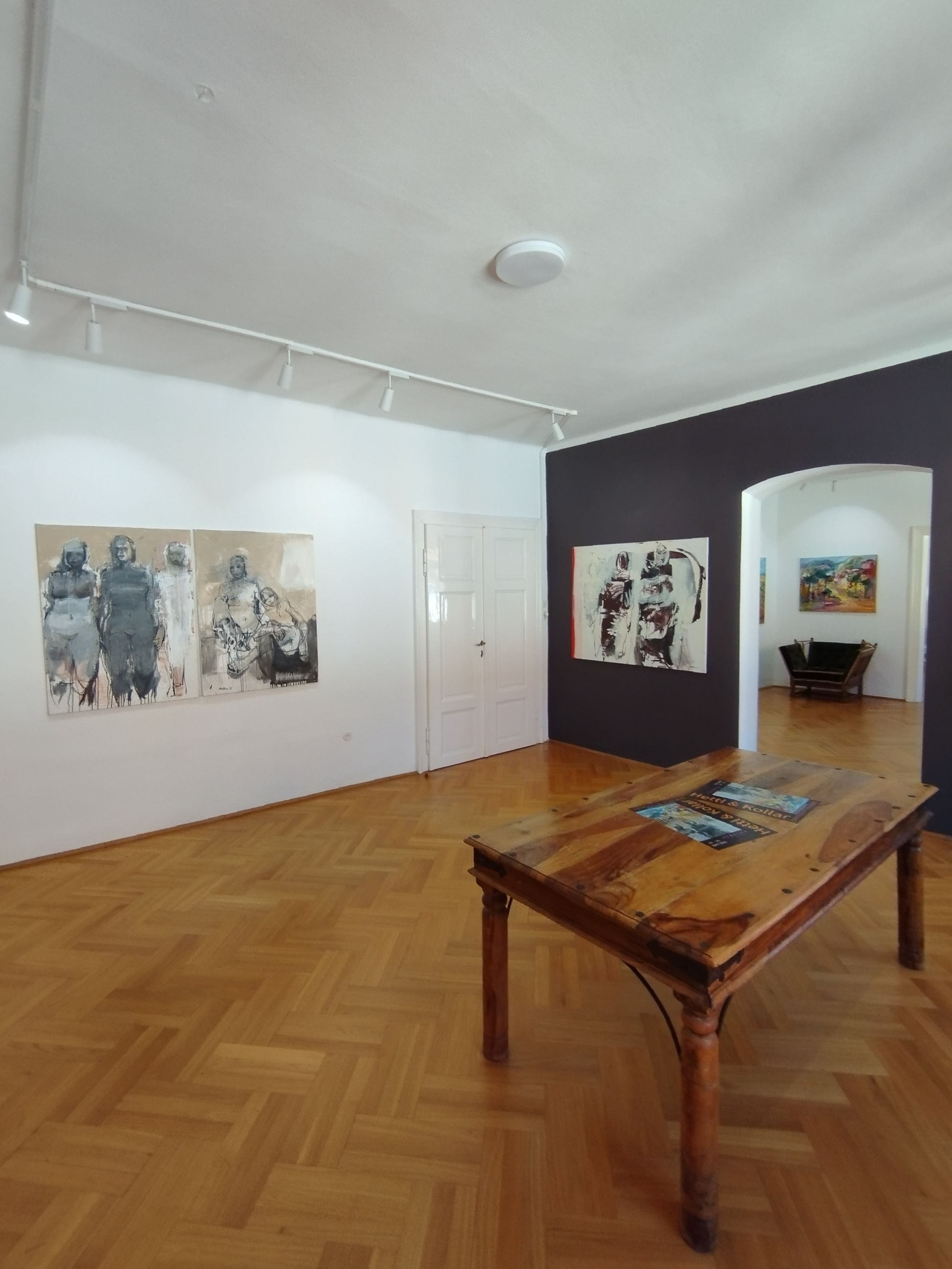 Innenansicht einer Kunstgalerie mit Holzboden, Gemälden an den Wänden und einem Holztisch in der Mitte.