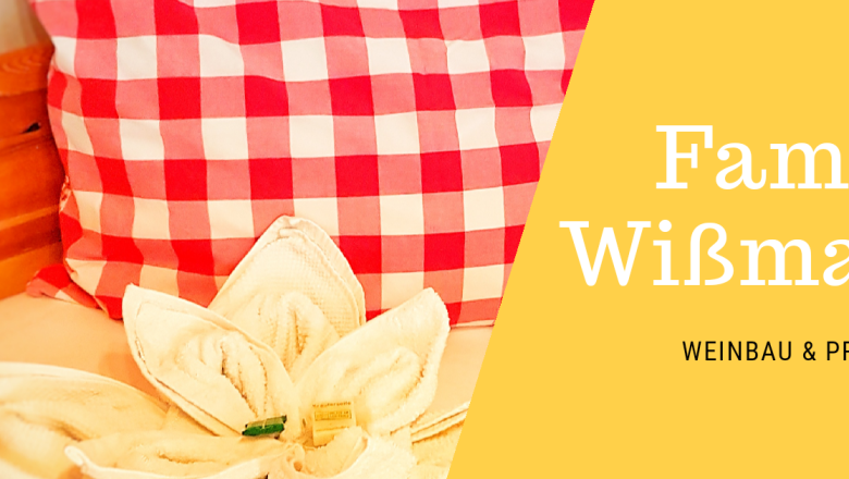 Rotes Kissen mit Karomuster und Handtuch in Blumenform auf einem Bett. Text: Familie Wißmann, Weinbau & Privatzimmer.
