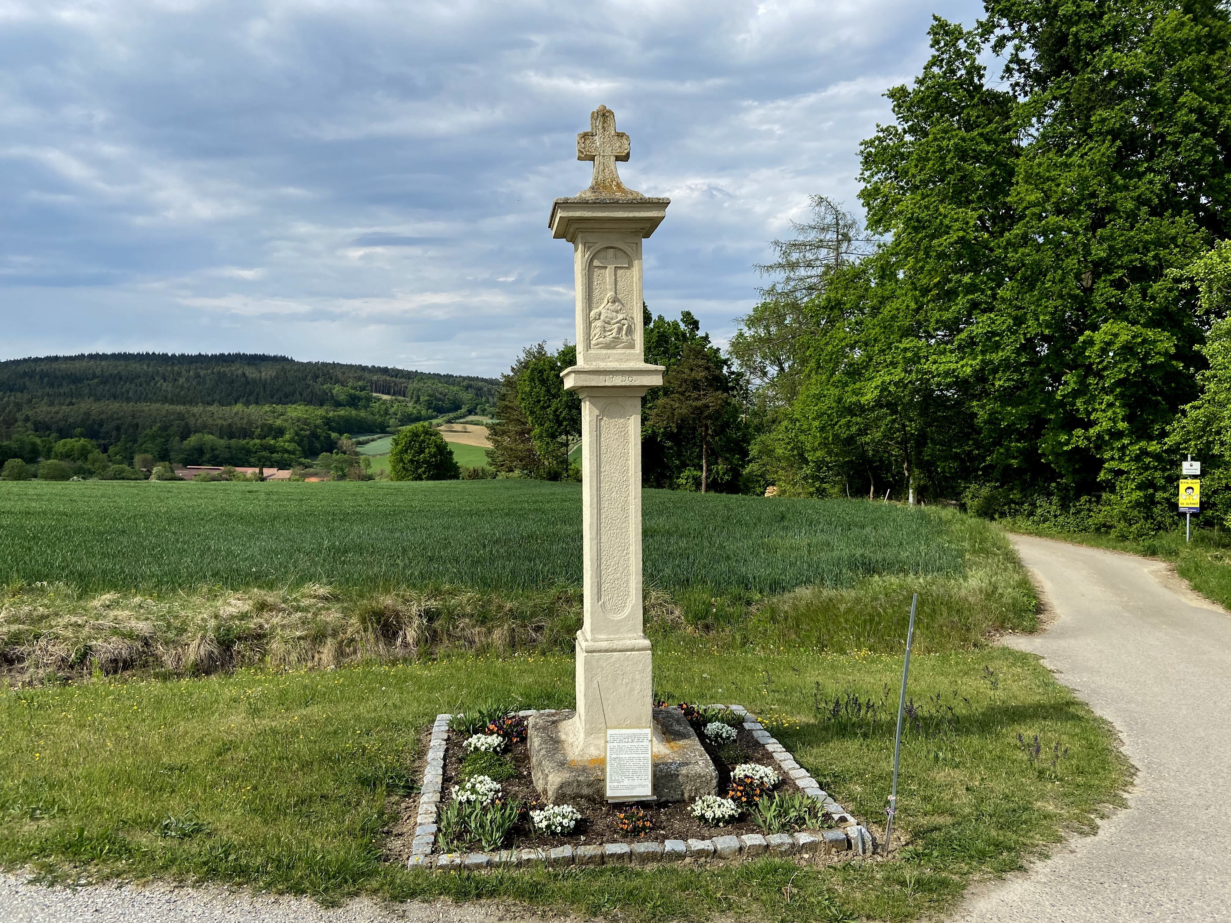 Steinkreuz auf einem Feldweg mit grüner Landschaft im Hintergrund.