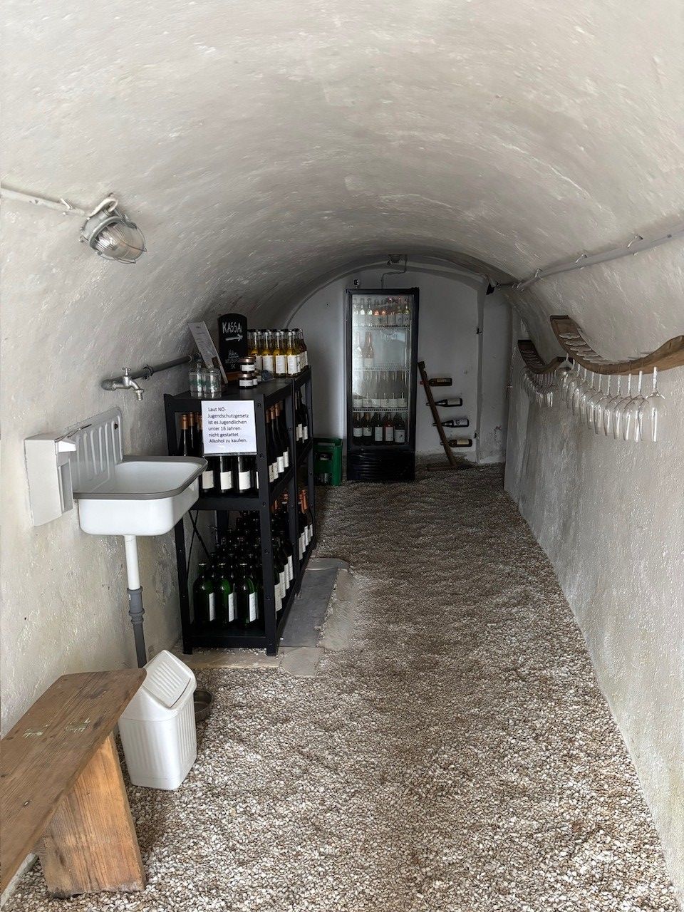 Innenraum einer schmalen Trinkstation im Gewölbekeller von Weinbau Ackerl mit Weinflaschen, Kühlschrank, Gläsern und Waschbecken.