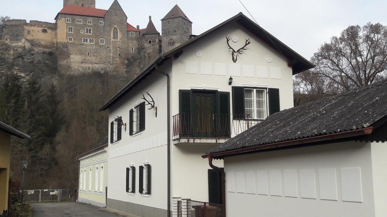 Ein weißes Haus mit grünen Fensterläden und Hirschgeweihen an der Wand, im Hintergrund eine Burg auf einem Hügel.