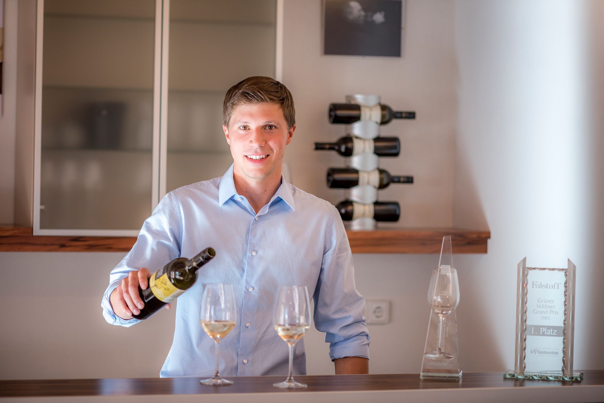 Ein junger Mann in einem blauen Hemd schenkt Wein in ein Glas ein, im Hintergrund Weinflaschen und eine Auszeichnung.