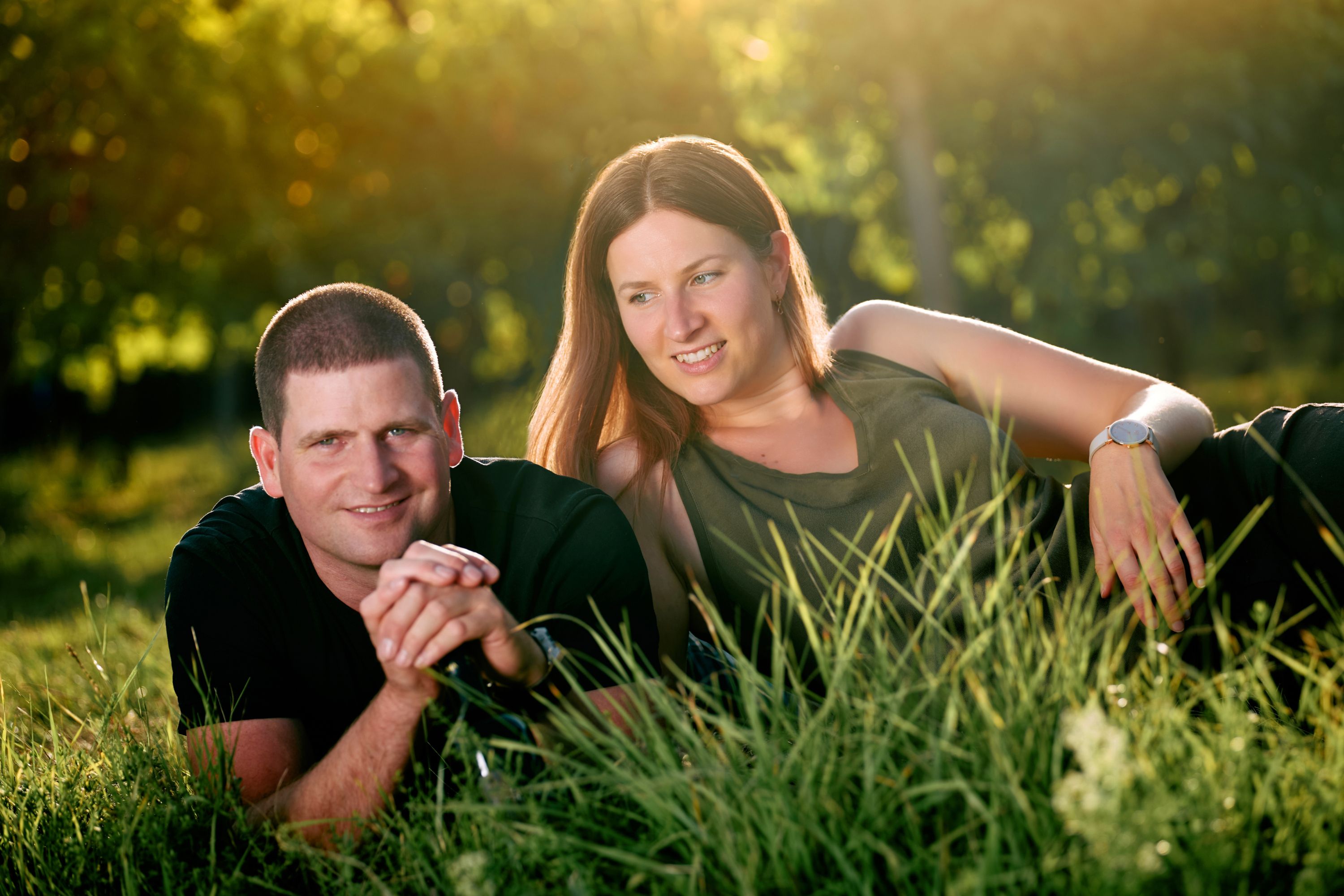 A man and a woman lie relaxed on the grass, surrounded by green nature and sunlight.