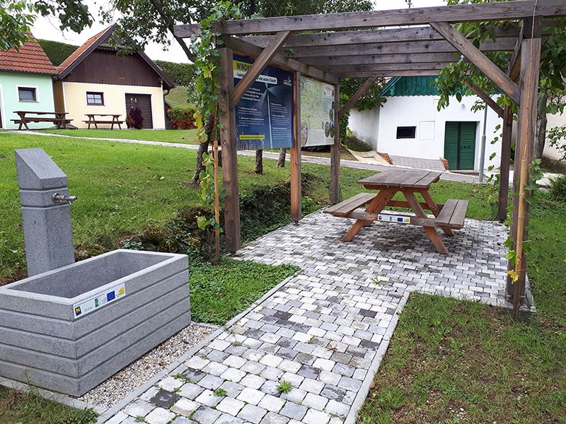 A rest area with a drinking fountain, wooden table and bench under a pergola, surrounded by green meadows and small buildings.