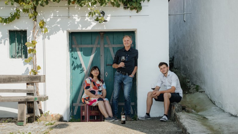 Three people in front of a wine cellar with a blue door and grapevines.