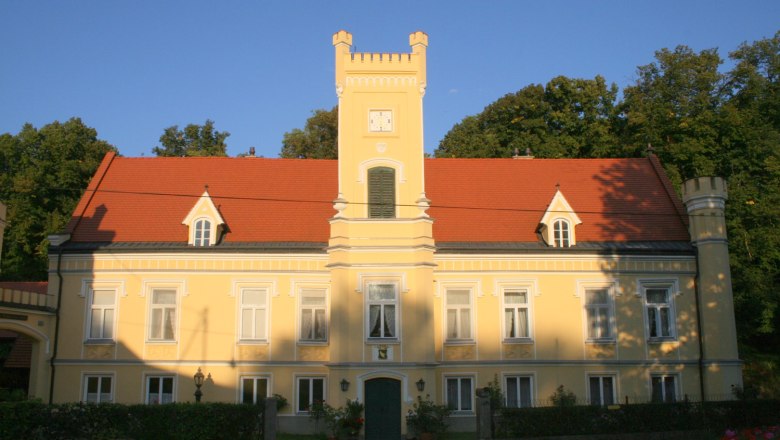 Gelbes Schloss mit rotem Dach und Turm, umgeben von Bäumen.