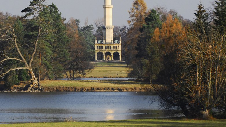 Ein Minarett im Schlosspark von Lednice, umgeben von Bäumen und einem See im Vordergrund.