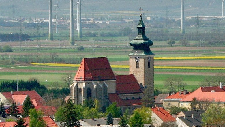 Kirche in Pillichsdorf mit umliegenden Häusern und Windrädern im Hintergrund.
