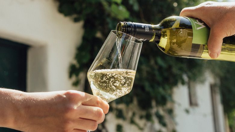 Eine Hand gießt Weißwein aus einer Flasche in ein Glas, das von einer anderen Hand gehalten wird.