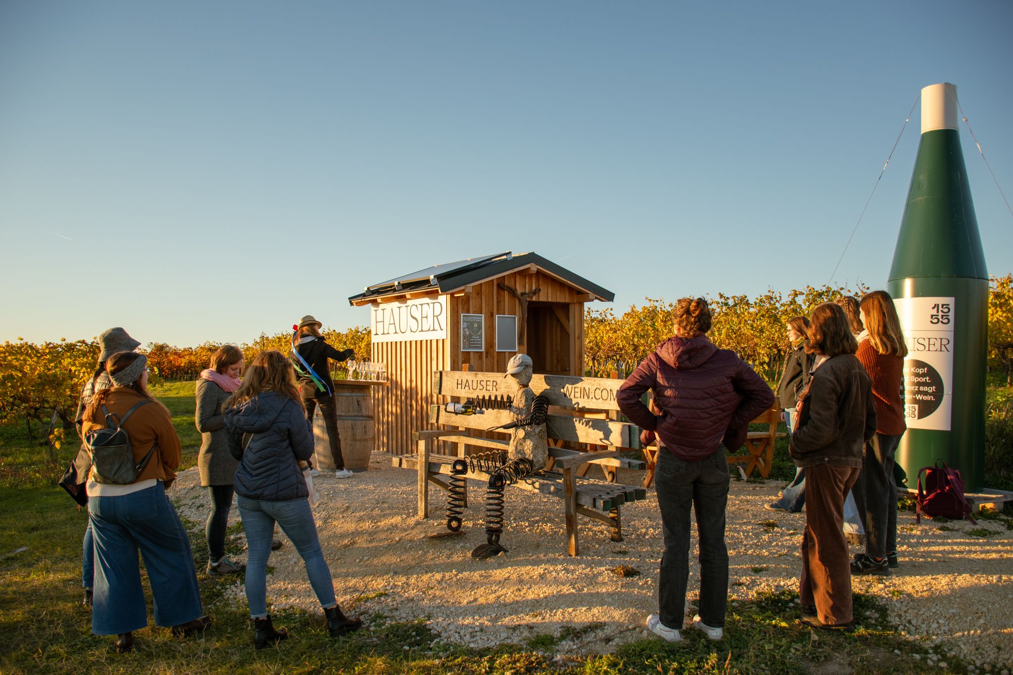 Gruppe von Menschen steht vor einem kleinen Holzhäuschen in einem Weinberg bei Sonnenuntergang.