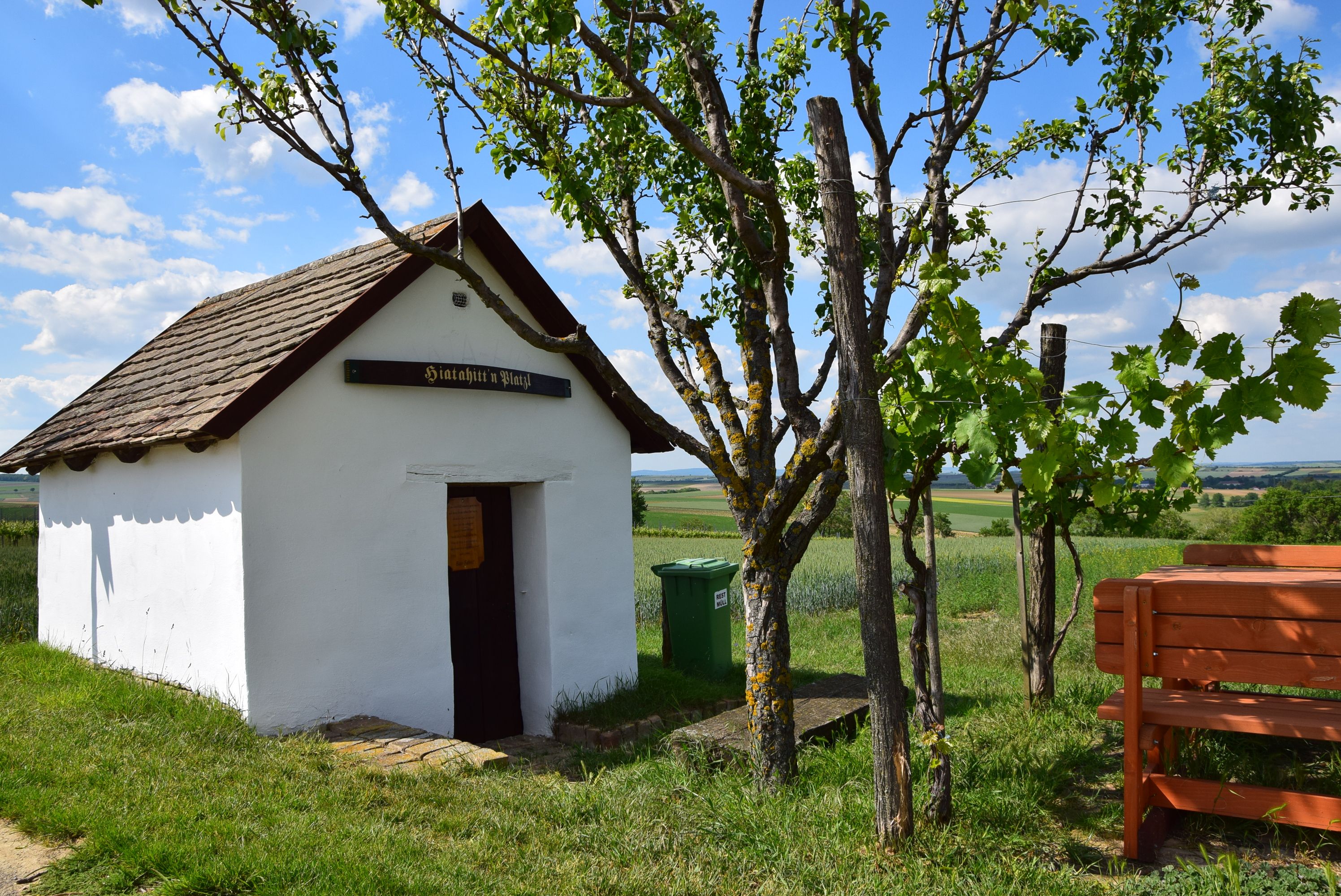 Kleine weiße Hütte mit Holzdach und Schild "Hiatahitt'n Platzl". Daneben ein Baum und eine Bank, im Hintergrund Felder und blauer Himmel.