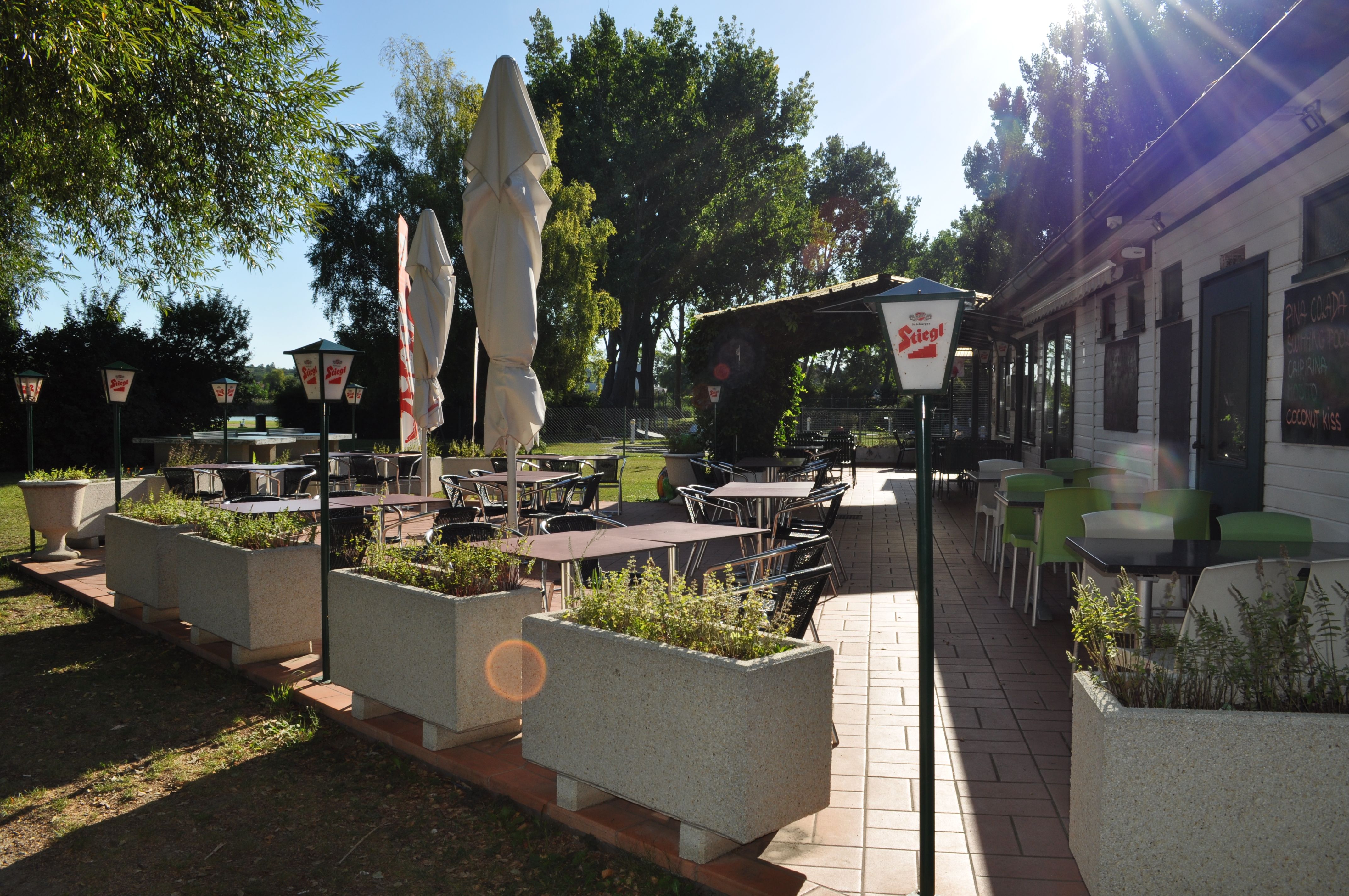 Sonnige Terrasse mit leeren Tischen und Stühlen, umgeben von Pflanzen und Sonnenschirmen.