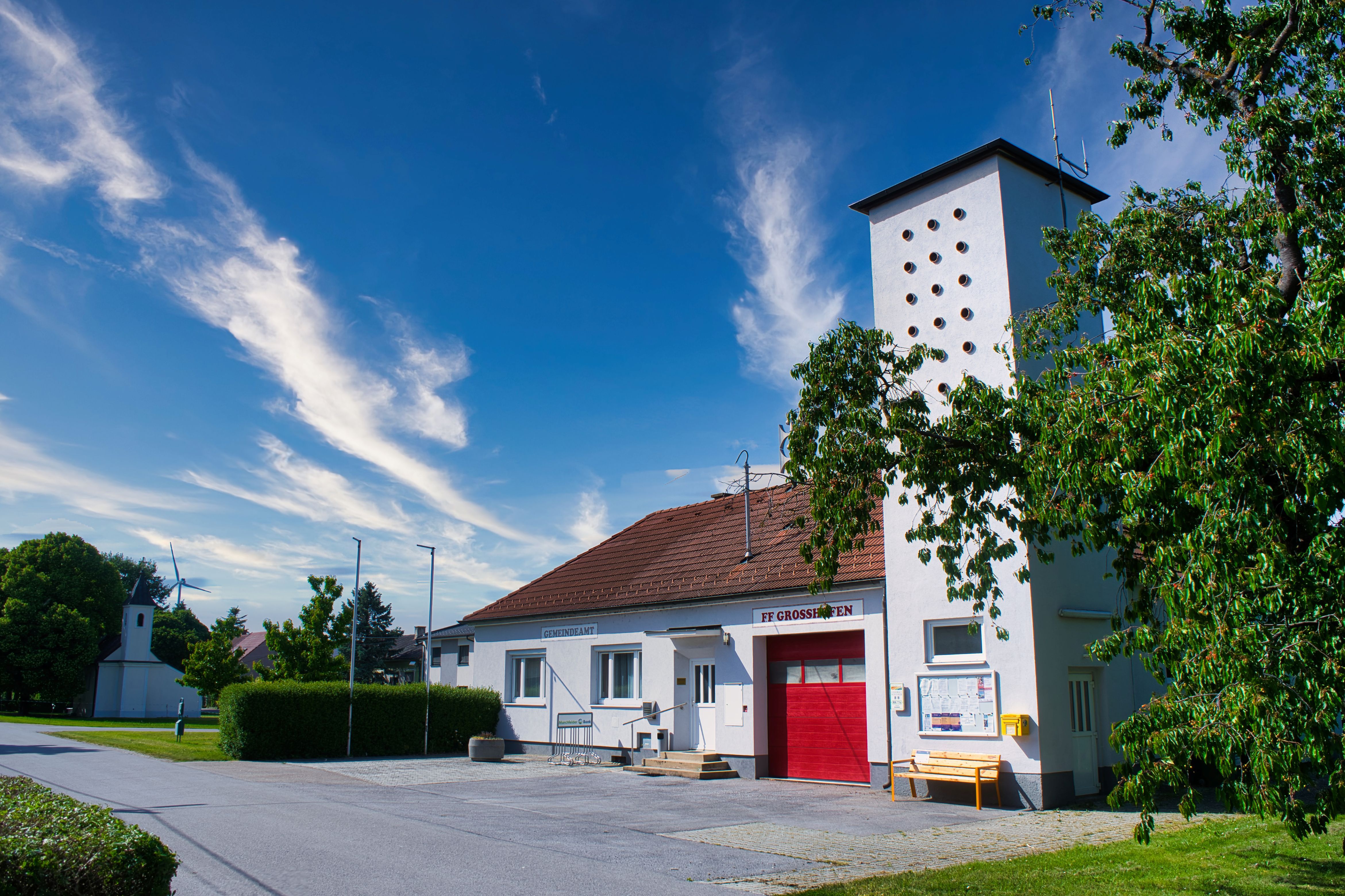 Gemeinde Großhofen mit Gemeindeamt und Feuerwehrhaus, weißem Turm, rotem Tor und Bäumen unter blauem Himmel.