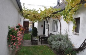 Ein gemütlicher Innenhof mit Holztisch und Bänken, umgeben von weißen Wänden, Blumen und Weinreben.