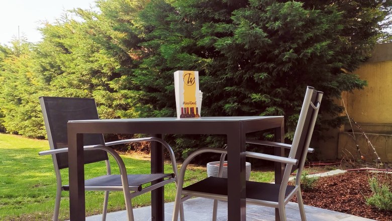 Terrasse mit Tisch, zwei Stühlen und Bäumen im Hintergrund.