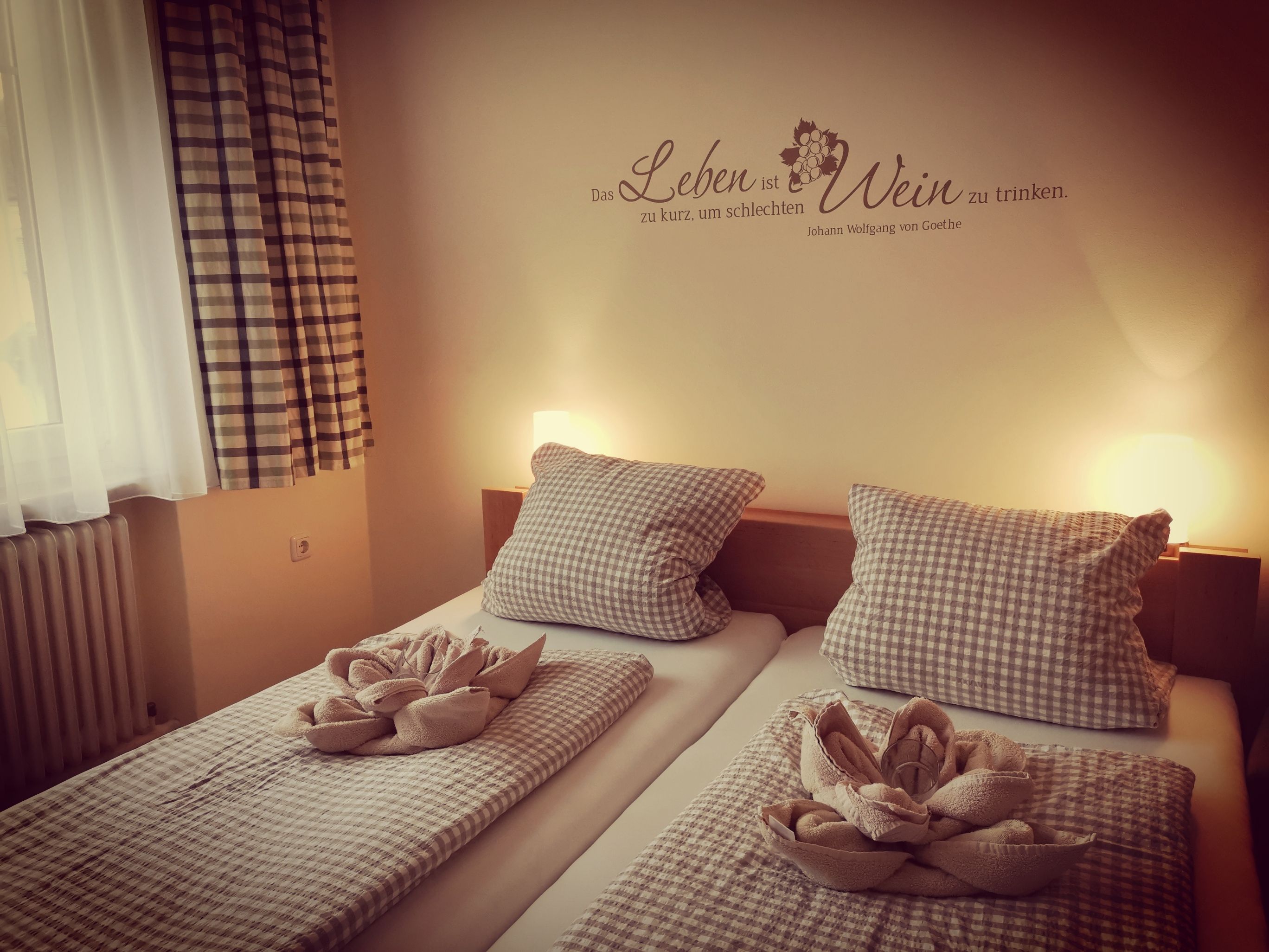 Gemütliches Gästezimmer mit zwei Betten, karierten Bettdecken und Kissen, dekorativen Handtüchern und einem Goethe-Zitat an der Wand.