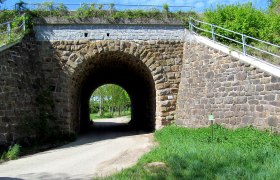 Steinviadukt mit Durchgang und grünem Umfeld.